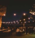 An image at night of the grass with tables for an event, market lights are overhead.